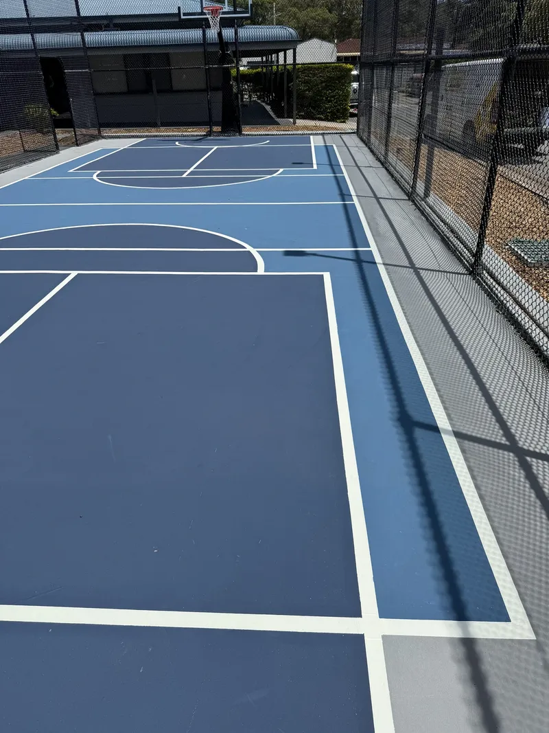 Long view down a freshly resurfaced two-tone blue school court in Sydney with crisp white line marking and perimeter fencing