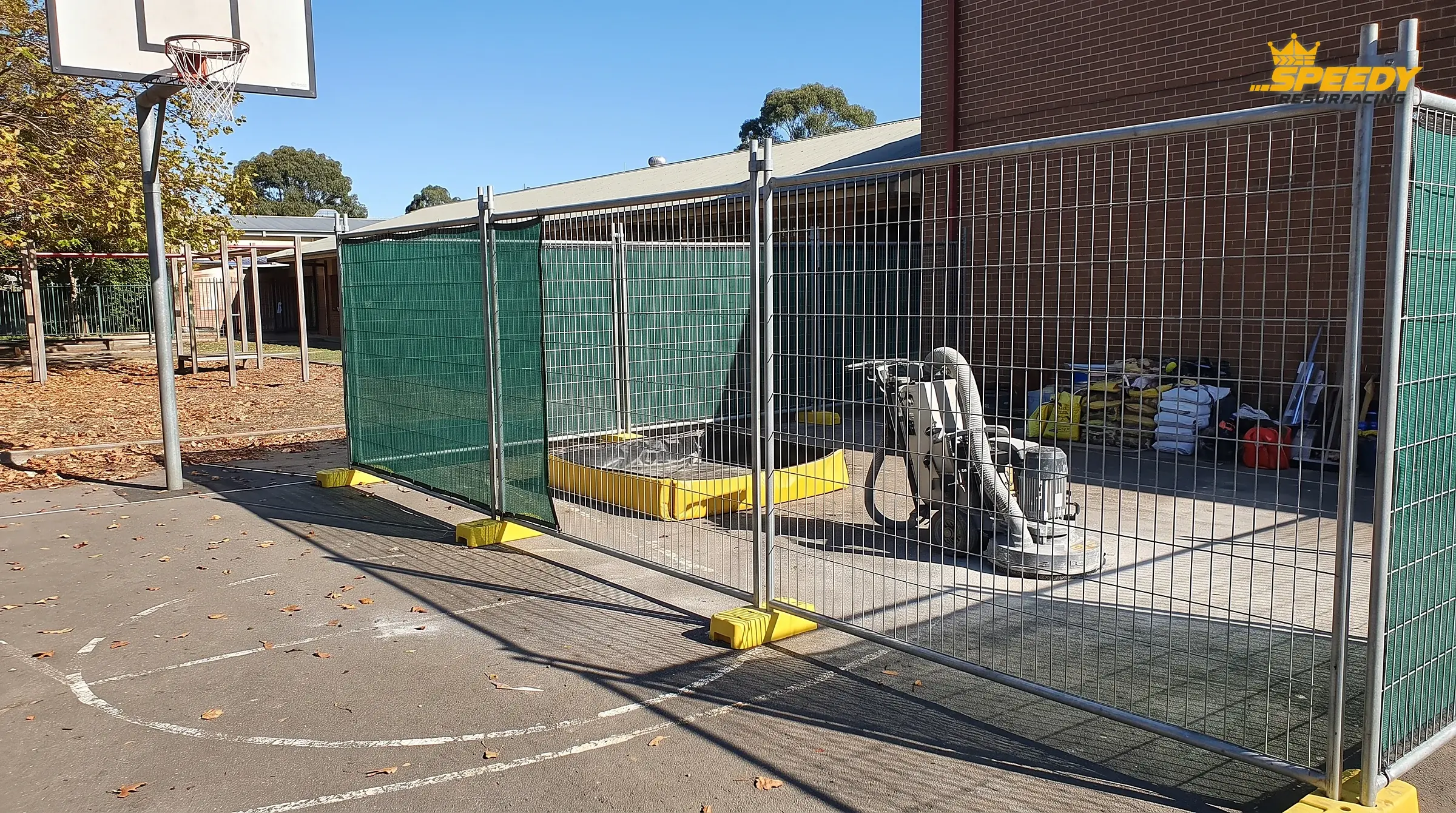 Structured site isolation: mesh fence panels, a vacuum-shrouded grinder and a wash-water bund keep the works contained while the adjacent playground stays open.