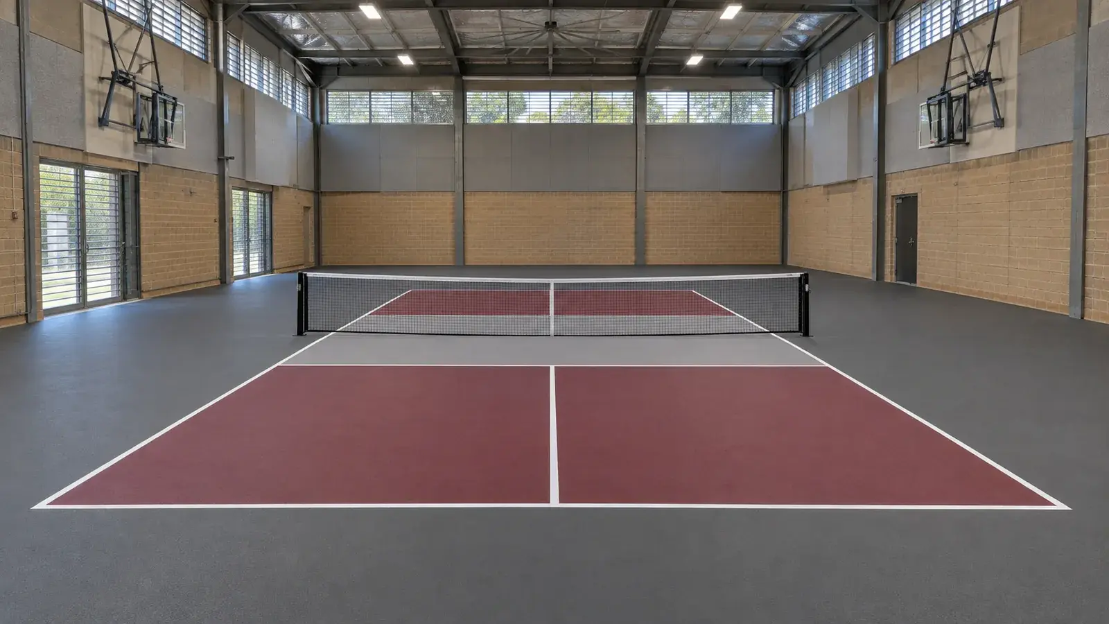 Two dedicated pickleball courts inside a NSW school hall — maroon acrylic play zones, grey run-offs and crisp white kitchen lines under basketball hoops.